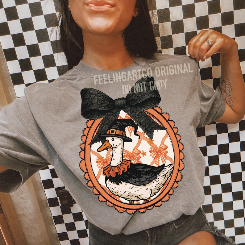 Peut inclure: T-shirt gris avec un motif d'Halloween. Le dessin repr&eacute;sente une oie portant un chapeau de sorci&egrave;re et un col &agrave; volants, dans un cadre ovale orange, avec un n&oelig;ud noir. Le texte "FEELINGARTCO ORIGINAL DO NOT COPY" est imprim&eacute; sur le t-shirt.