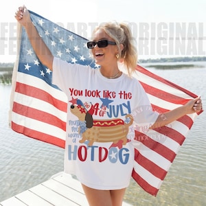 May include: White t-shirt with a graphic of a hotdog-shaped dog wearing a patriotic hat and sunglasses. The shirt has the text "You look like the 4th of July" and "makes me want a HOTDOG". The person is holding an American flag.