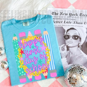 Puede incluir: Una camiseta azul claro con un gr&aacute;fico de l&aacute;pices coloridos y un marco festoneado amarillo que rodea el texto rosa "happy first day of school". El dise&ntilde;o incluye peque&ntilde;as estrellas y se muestra junto a un peri&oacute;dico y bolas de discoteca.