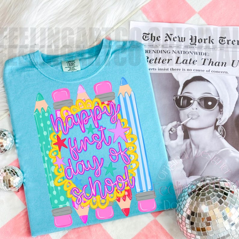 Puede incluir: Una camiseta azul claro con un gr&aacute;fico de l&aacute;pices coloridos y un marco festoneado amarillo que rodea el texto rosa "happy first day of school". El dise&ntilde;o incluye peque&ntilde;as estrellas y se muestra junto a un peri&oacute;dico y bolas de discoteca.
