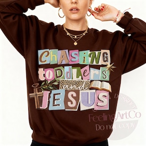 Pu&ograve; includere: Una felpa marrone con la scritta "Chasing Toddlers and Jesus" in uno stile colorato e ritagliato. Il design include una croce, un libro aperto e una stella. La felpa &egrave; uno stile comodo e casual.