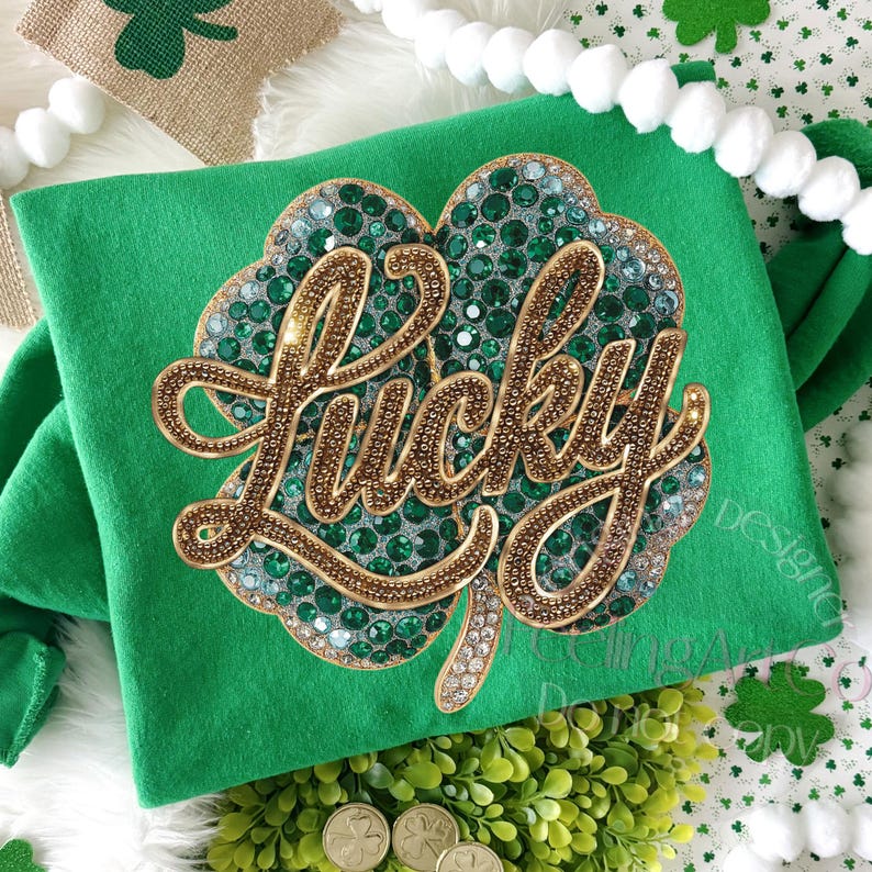 May include: Green t-shirt with a large, sparkling shamrock design. The shamrock is embellished with green and clear rhinestones and outlined in gold. The word "LUCKY" is written in gold script across the shamrock.