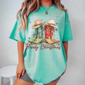 May include: A turquoise t-shirt features a graphic of two cowboy boots and hats decorated with Christmas lights. The boots are green and red, and the hats are tan. The text "Howdy Christmas" is written in red script below the boots.