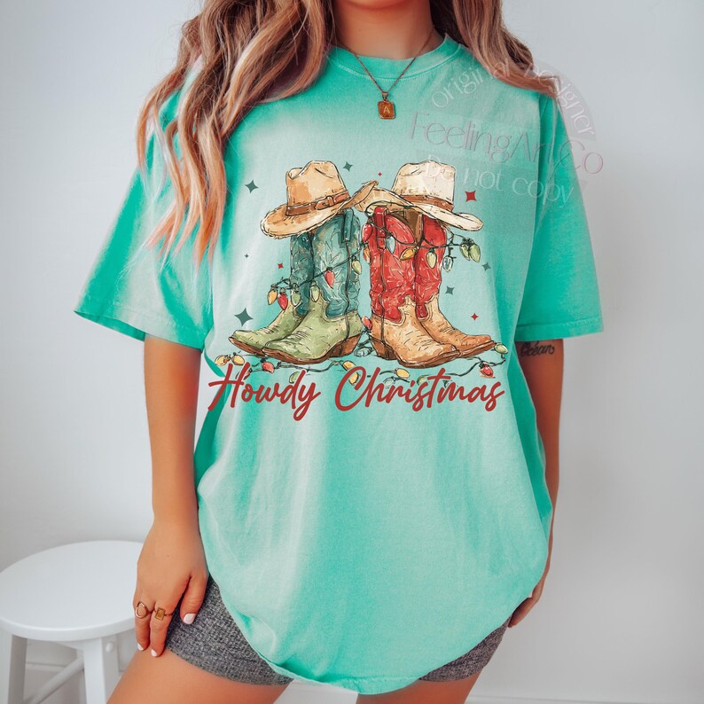 May include: A turquoise t-shirt features a graphic of two cowboy boots and hats decorated with Christmas lights. The boots are green and red, and the hats are tan. The text "Howdy Christmas" is written in red script below the boots.