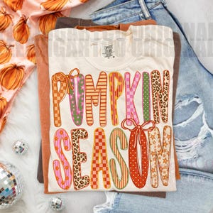 May include: A cream-colored t-shirt featuring the text PUMPKIN SEASON in large, multi-patterned letters with orange, pink, green, and brown designs, including leopard print, checkers, and stars, with two orange bows accenting the lettering.
