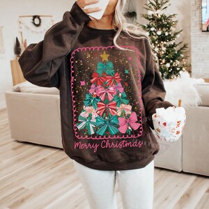 May include: A dark brown sweatshirt with a Christmas tree design composed of colourful bows. The tree is framed with a pink border and a gold star. The words "Merry Christmas" are written in pink script at the bottom of the design.