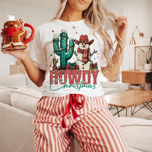 Peut inclure: T-shirt blanc avec un motif festif repr&eacute;sentant un cactus, un bonhomme de neige et l'inscription "HOWDY Christmas". Le bonhomme de neige porte un chapeau de cowboy et une &eacute;charpe. La personne porte un pantalon de pyjama &agrave; rayures rouges et blanches.