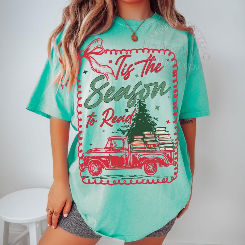 May include: A turquoise t-shirt with a red graphic of a vintage truck loaded with books and a Christmas tree. The text reads "Tis The Season to Read" in a festive font, framed by a red border and a red bow.