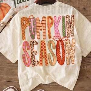 May include: A cream-colored short-sleeve t-shirt featuring the text PUMPKIN SEASON in large, multi-patterned letters. The design includes orange, pink, and green colors with checkered, leopard print, star, and striped patterns, accented by bows.
