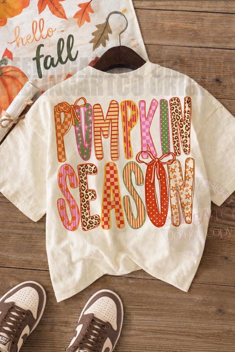 May include: A cream-colored short-sleeve t-shirt featuring the text PUMPKIN SEASON in large, multi-patterned letters. The design includes orange, pink, and green colors with checkered, leopard print, star, and striped patterns, accented by bows.