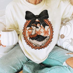 Peut inclure: T-shirt cr&egrave;me avec un dessin d'oie de dessin anim&eacute; portant un chapeau de p&egrave;lerin et un col &agrave; volants, dans un cadre ovale orange. Un grand n&oelig;ud noir surmonte le motif. Le t-shirt est port&eacute; avec un jean bleu clair.