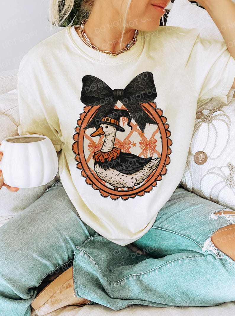 Peut inclure: T-shirt cr&egrave;me avec un dessin d'oie de dessin anim&eacute; portant un chapeau de p&egrave;lerin et un col &agrave; volants, dans un cadre ovale orange. Un grand n&oelig;ud noir surmonte le motif. Le t-shirt est port&eacute; avec un jean bleu clair.