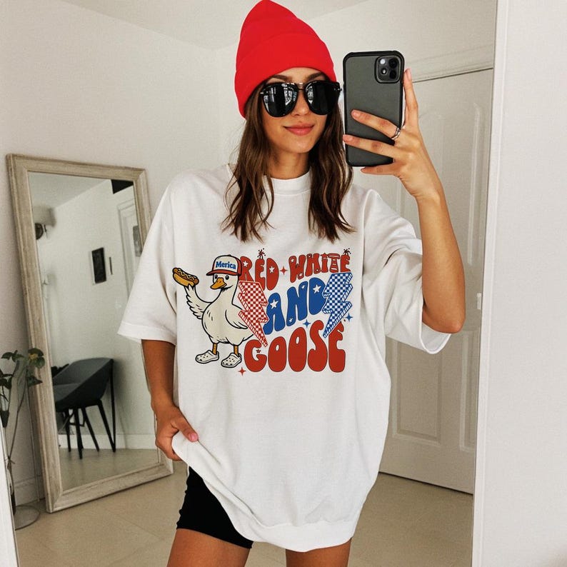 May include: White t-shirt featuring a cartoon goose with a hot dog and a hat that says "Merica". The shirt has the text "Red, White, and Goose" in red, white, and blue. The person in the image is wearing a red beanie and sunglasses.
