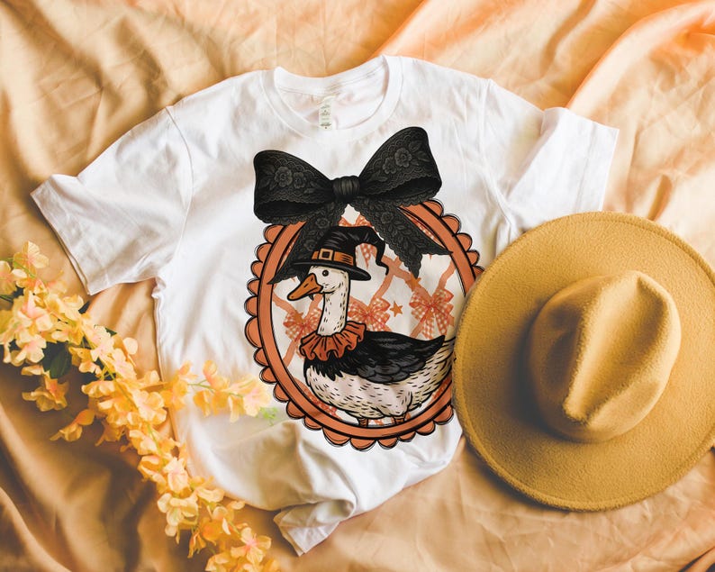 Peut inclure: T-shirt blanc avec un dessin d'oie de dessin anim&eacute; portant un chapeau de sorci&egrave;re et un col &agrave; volants, encadr&eacute; d'un n&oelig;ud noir. Un chapeau jaune et des fleurs sont &agrave; c&ocirc;t&eacute; du t-shirt. Le design est sur le th&egrave;me d'Halloween.