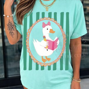 May include: A light teal t-shirt featuring a cartoon goose wearing a pink bow and reading a pink book, holding a pencil. The goose is inside a circular frame with a scalloped edge. Vertical green stripes are on the shirt.