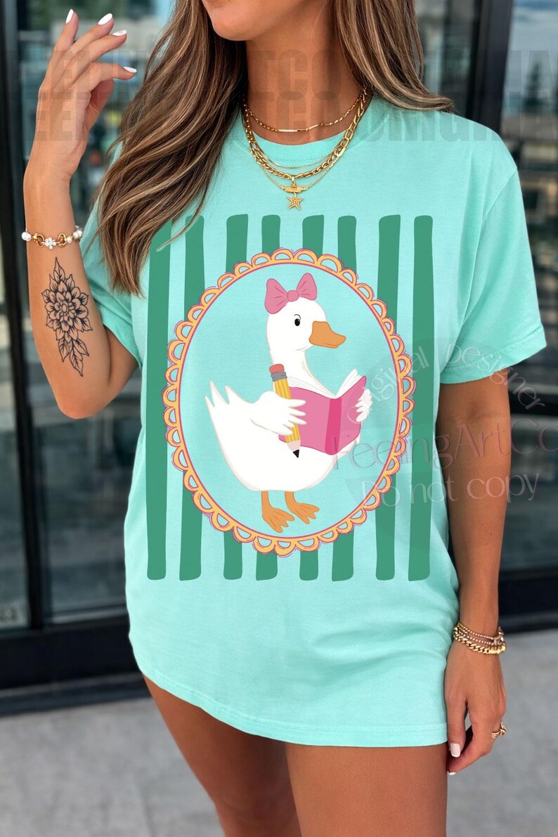 May include: A light teal t-shirt featuring a cartoon goose wearing a pink bow and reading a pink book, holding a pencil. The goose is inside a circular frame with a scalloped edge. Vertical green stripes are on the shirt.