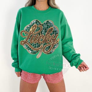 May include: A green sweatshirt with a large, sequined shamrock design. The word "Lucky" is embroidered in gold script across the shamrock. The model is wearing red and white checkered shorts.
