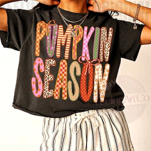 May include: A black short-sleeved t-shirt featuring the text PUMPKIN SEASON in large, multi-colored letters with various patterns including leopard print, checkers, stars, and stripes, paired with white and black vertical striped shorts.