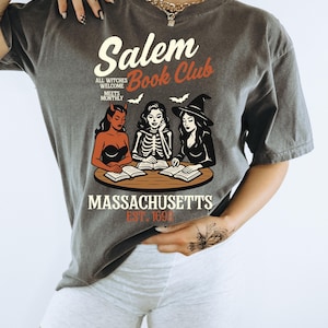 May include: Dark gray t-shirt with a Salem Book Club graphic. The design features three witches, books, and the text "Massachusetts Est. 1692". The shirt has a vintage, slightly faded look.