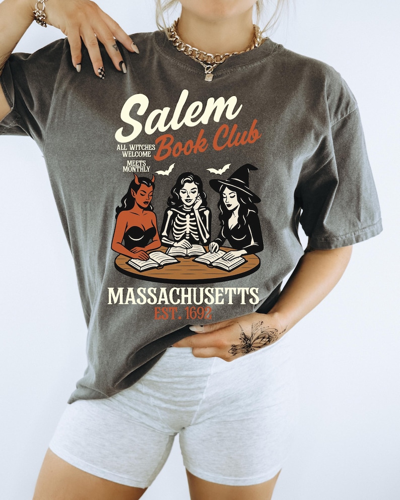 May include: Dark gray t-shirt with a Salem Book Club graphic. The design features three witches, books, and the text "Massachusetts Est. 1692". The shirt has a vintage, slightly faded look.