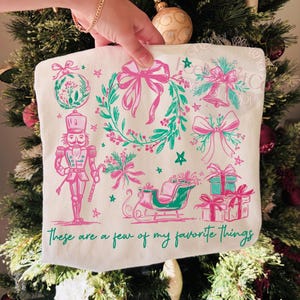 May include: A white tote bag featuring a Christmas-themed design in pink and teal. The design includes a wreath, bows, a bell, a nutcracker, a sleigh, and gifts. The text "These are a few of my favorite things" is printed at the bottom.