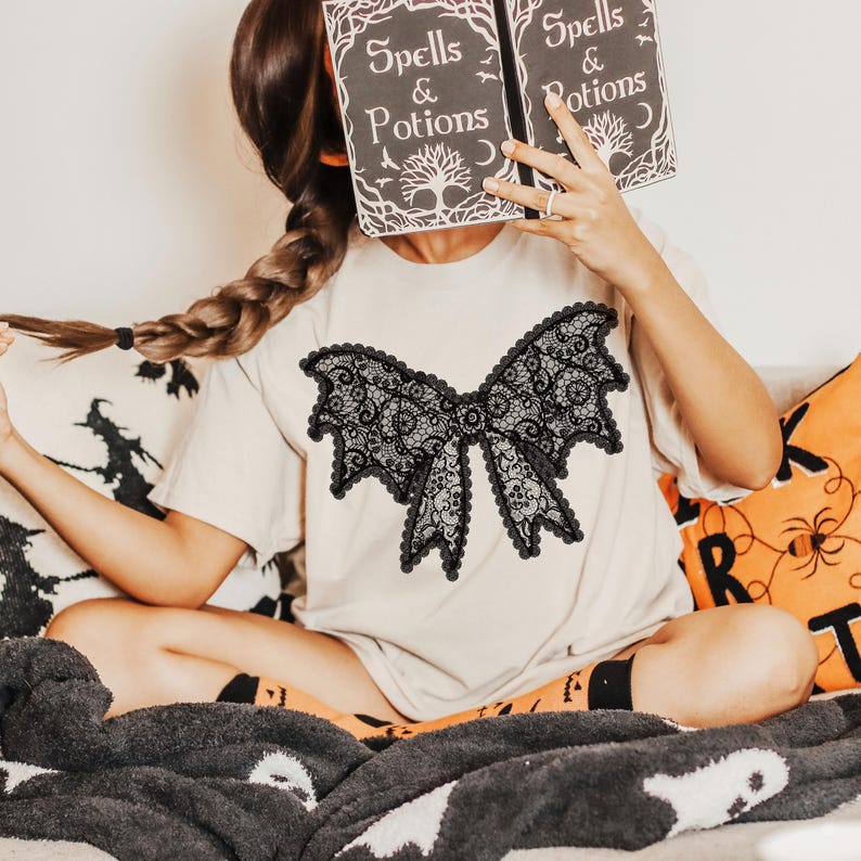 May include: A beige t-shirt featuring a large black lace bow design. The person in the image is holding a book with the words "Spells & Potions" on the cover. The image has a Halloween theme.