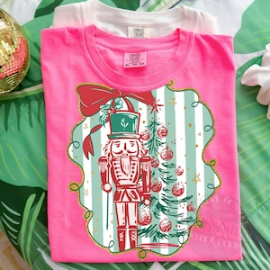 May include: A vibrant pink t-shirt with a festive Christmas design. The graphic includes a red and white nutcracker, a Christmas tree, and a red bow. The shirt has a crew neck and short sleeves. A white t-shirt is behind the pink one.