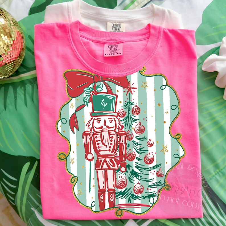 May include: A vibrant pink t-shirt with a festive Christmas design. The graphic includes a red and white nutcracker, a Christmas tree, and a red bow. The shirt has a crew neck and short sleeves. A white t-shirt is behind the pink one.