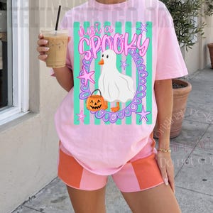 May include: A light pink t-shirt featuring a graphic of a white ghost-costumed duck holding an orange jack-o'-lantern, surrounded by a purple oval frame and green vertical stripes, with the text Just a SPOOKY printed in pink above.