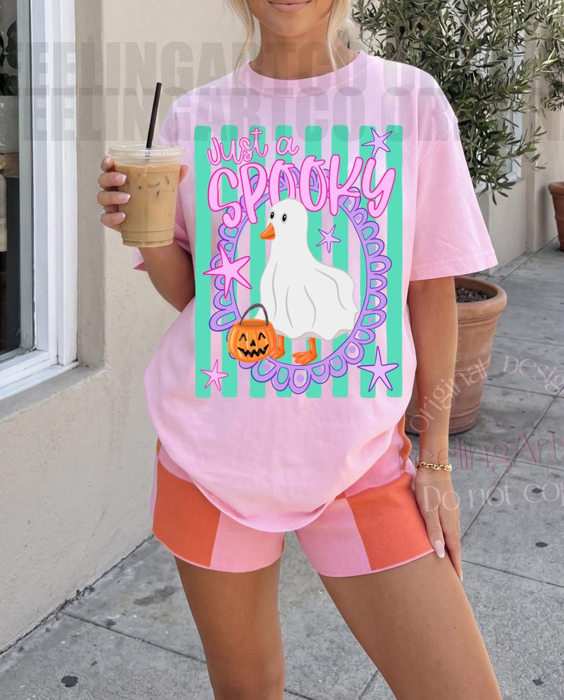 May include: A light pink t-shirt featuring a graphic of a white ghost-costumed duck holding an orange jack-o'-lantern, surrounded by a purple oval frame and green vertical stripes, with the text Just a SPOOKY printed in pink above.