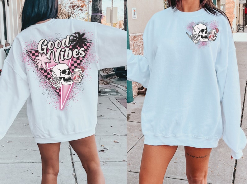 Puede incluir: Una sudadera blanca con un dise&ntilde;o gr&aacute;fico rosa y negro en la espalda. El gr&aacute;fico presenta una calavera con una bebida, palmeras y el texto "Good Vibes". La parte delantera de la sudadera es blanca lisa.