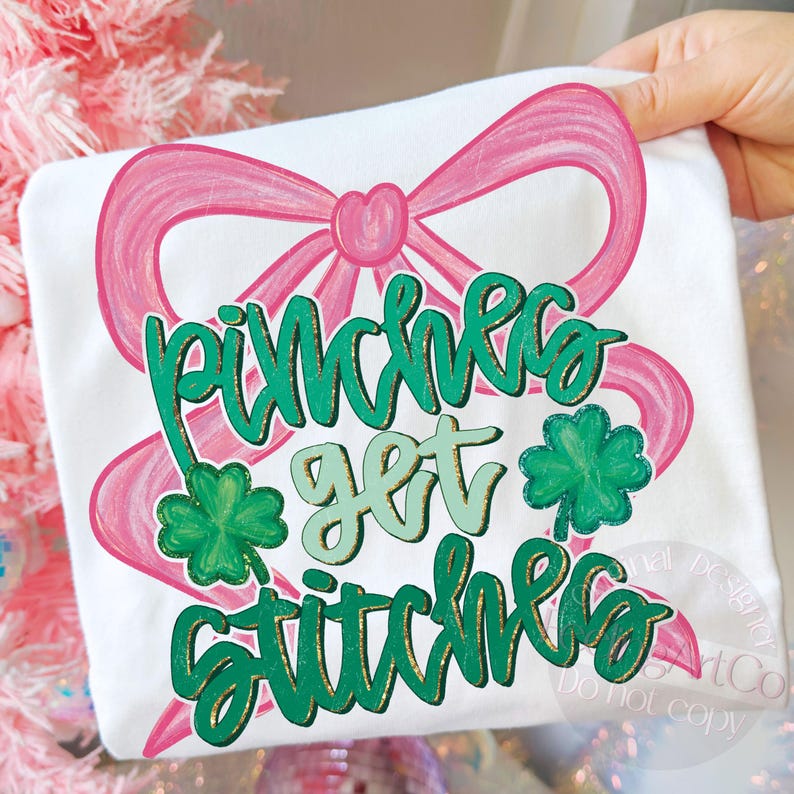 May include: White t-shirt with a pink bow and the text "Pinches Get Stitches" in green and gold lettering. Two green shamrocks are also featured on the shirt. The design is likely for St. Patrick's Day.
