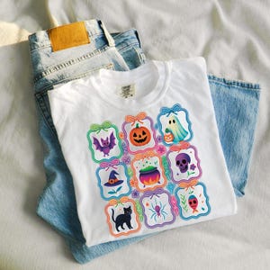 May include: White t-shirt with colorful Halloween-themed graphics, including a pumpkin, ghost, bat, and witch's hat. The shirt is folded on top of a pair of light blue denim jeans.