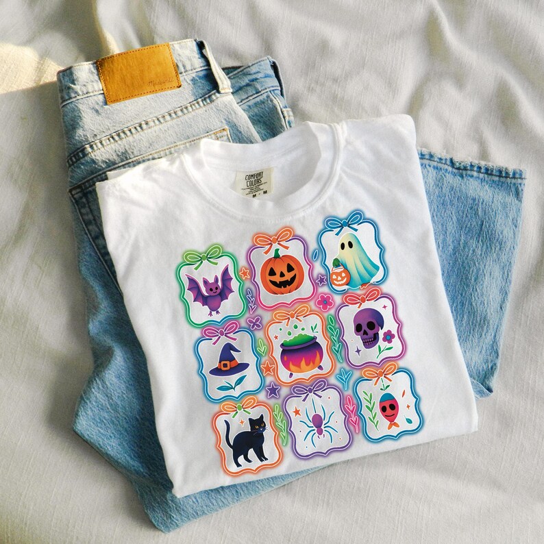 May include: White t-shirt with colorful Halloween-themed graphics, including a pumpkin, ghost, bat, and witch's hat. The shirt is folded on top of a pair of light blue denim jeans.