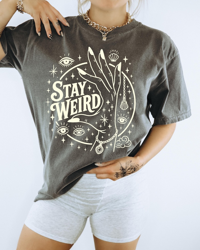 May include: Dark gray t-shirt with the words "STAY WEIRD" in a circular design. The design includes a hand, eyes, stars, and a pendant. The shirt is a short-sleeved, casual style.