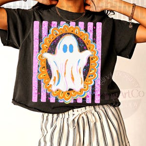 May include: A black short-sleeve t-shirt featuring a graphic print of a white ghost with blue eyes centred inside an orange scalloped frame. The design is set against a background of vertical pink stripes with blue and pink paint splatter accents.