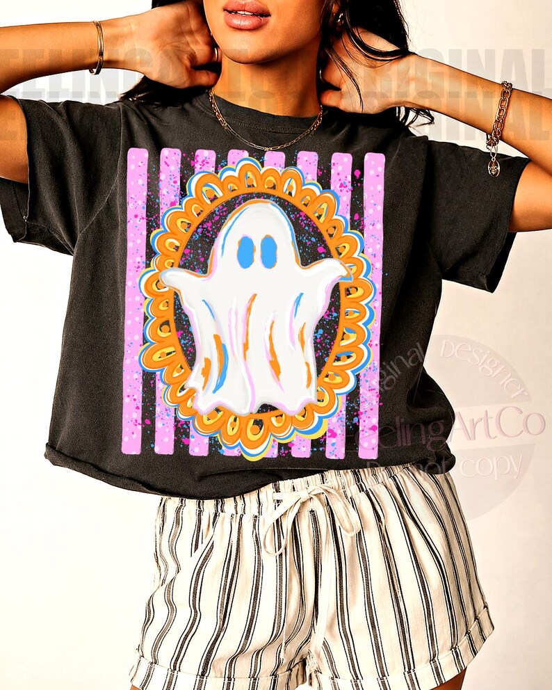 May include: A black short-sleeve t-shirt featuring a graphic print of a white ghost with blue eyes centred inside an orange scalloped frame. The design is set against a background of vertical pink stripes with blue and pink paint splatter accents.