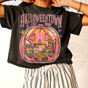 May include: A black short-sleeve t-shirt featuring a large orange pumpkin graphic with a carved face. Inside the pumpkin is a scene of a house and a graveyard. The text reads HALLOWEENTOWN EST. 1998 in blue and pink letters with small stars.