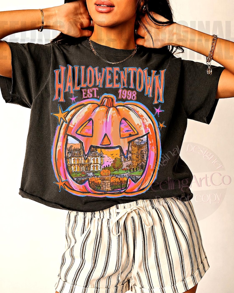 May include: A black short-sleeve t-shirt featuring a large orange pumpkin graphic with a carved face. Inside the pumpkin is a scene of a house and a graveyard. The text reads HALLOWEENTOWN EST. 1998 in blue and pink letters with small stars.