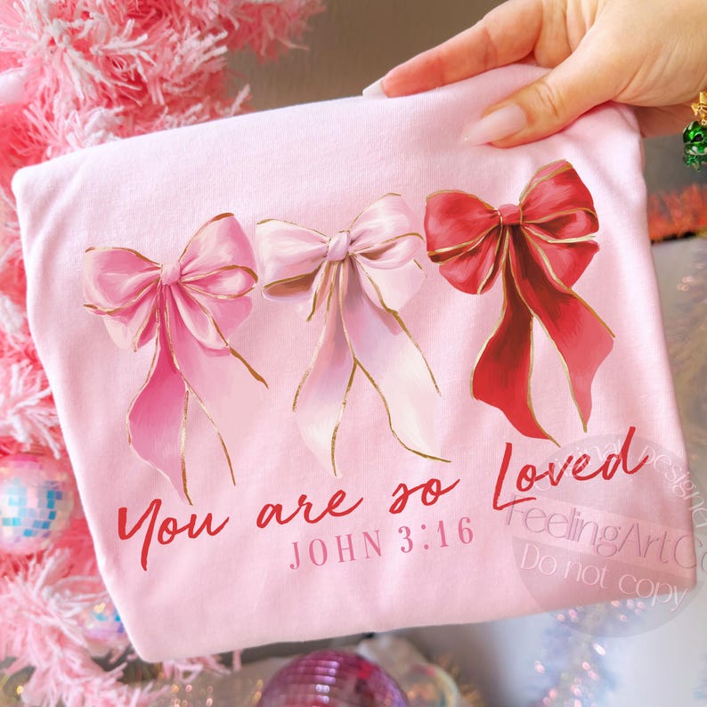 Pu&ograve; includere: T-shirt rosa chiaro con tre fiocchi decorativi rosa e rossi con bordi dorati. Il testo "You are so Loved" e "JOHN 3:16" &egrave; stampato in rosso sulla maglietta.