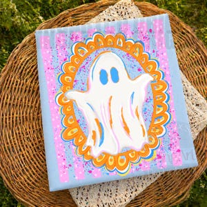 May include: A square canvas print featuring a central white ghost with blue eyes, enclosed within an orange and yellow scalloped oval frame. The background consists of light blue vertical stripes with pink and blue paint splatter accents.