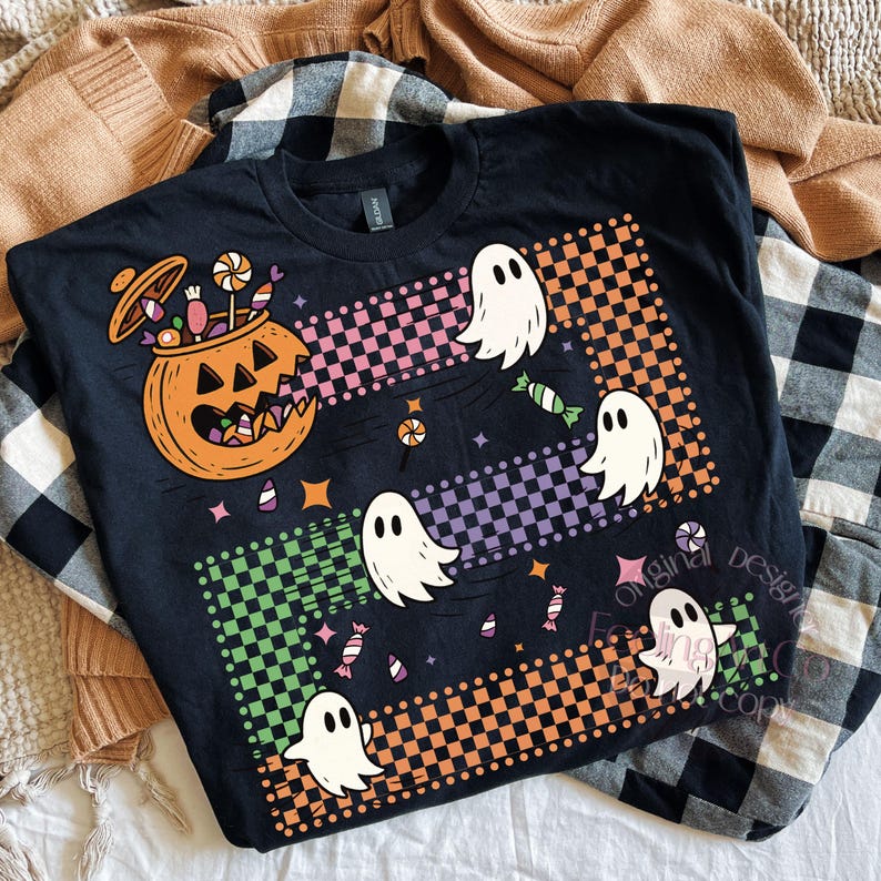 May include: Black t-shirt featuring a Halloween design. The graphic includes a jack-o'-lantern, ghosts, and candy. The design incorporates checkered patterns in orange, pink, green, and purple. The shirt is displayed on a neutral background.