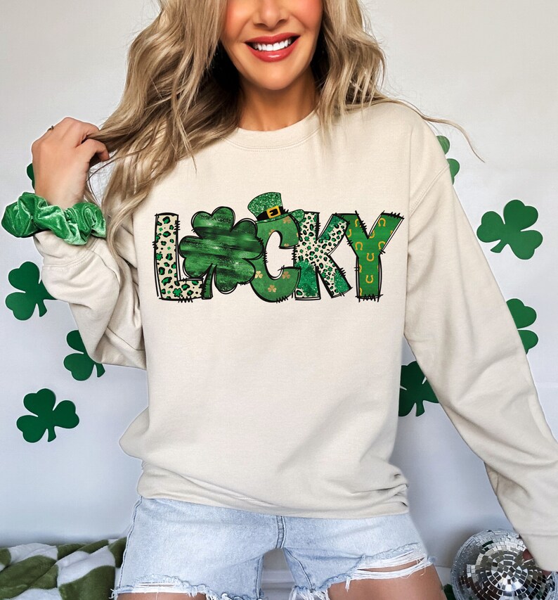 May include: A beige sweatshirt with the word "LUCKY" in a green and leopard print design. The letter "O" is a shamrock. The sweatshirt is long-sleeved and has a crew neck.