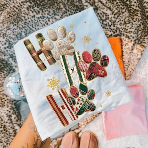 K&ouml;nnte beinhalten: Ein hellblaues, weihnachtliches Dekotuch mit dem gestickten Text "HO HO HO" und Pfotenabdr&uuml;cken. Die Buchstaben und Pfotenabdr&uuml;cke sind mit Karomustern und festlichen Mustern verziert. Goldene Schneeflocken sind verstreut.
