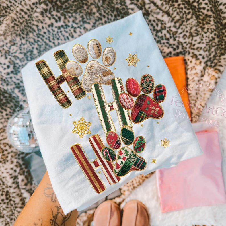 K&ouml;nnte beinhalten: Ein hellblaues, weihnachtliches Dekotuch mit dem gestickten Text "HO HO HO" und Pfotenabdr&uuml;cken. Die Buchstaben und Pfotenabdr&uuml;cke sind mit Karomustern und festlichen Mustern verziert. Goldene Schneeflocken sind verstreut.