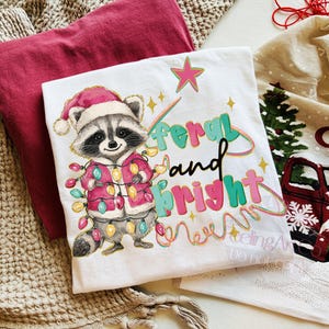May include: White t-shirt featuring a raccoon wearing a Santa hat and a pink coat, adorned with Christmas lights. The text "feral and bright" is printed in colorful letters. A red t-shirt is visible in the background.