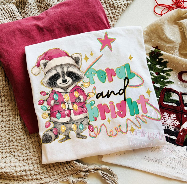 May include: White t-shirt featuring a raccoon wearing a Santa hat and a pink coat, adorned with Christmas lights. The text "feral and bright" is printed in colorful letters. A red t-shirt is visible in the background.