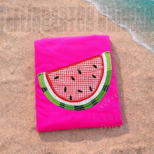 May include: A bright pink t-shirt featuring a watermelon slice applique. The watermelon design includes a red and white checkered pattern, green rind, and black seeds. The shirt is lying on a sandy beach.