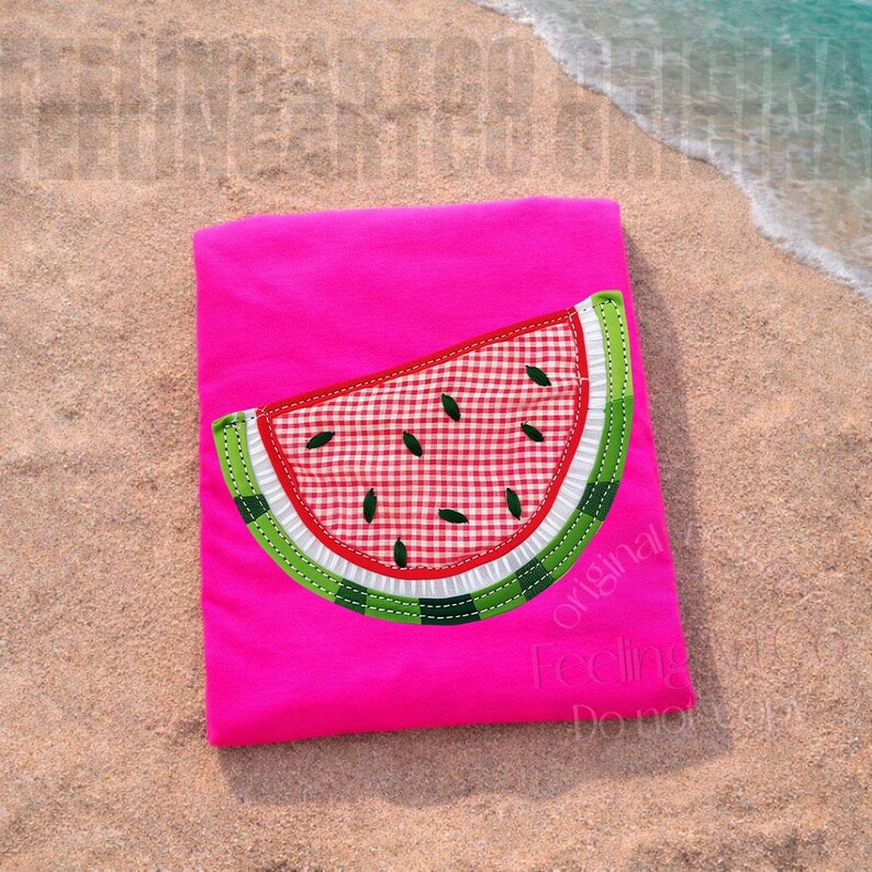 May include: A bright pink t-shirt featuring a watermelon slice applique. The watermelon design includes a red and white checkered pattern, green rind, and black seeds. The shirt is lying on a sandy beach.