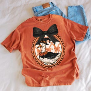 Peut inclure: T-shirt orange avec un dessin d'oie de dessin anim&eacute; portant un chapeau de sorci&egrave;re et un col &agrave; volants, encadr&eacute; d'un n&oelig;ud noir. Le motif est sur un t-shirt orange clair, avec un jean bleu en arri&egrave;re-plan.